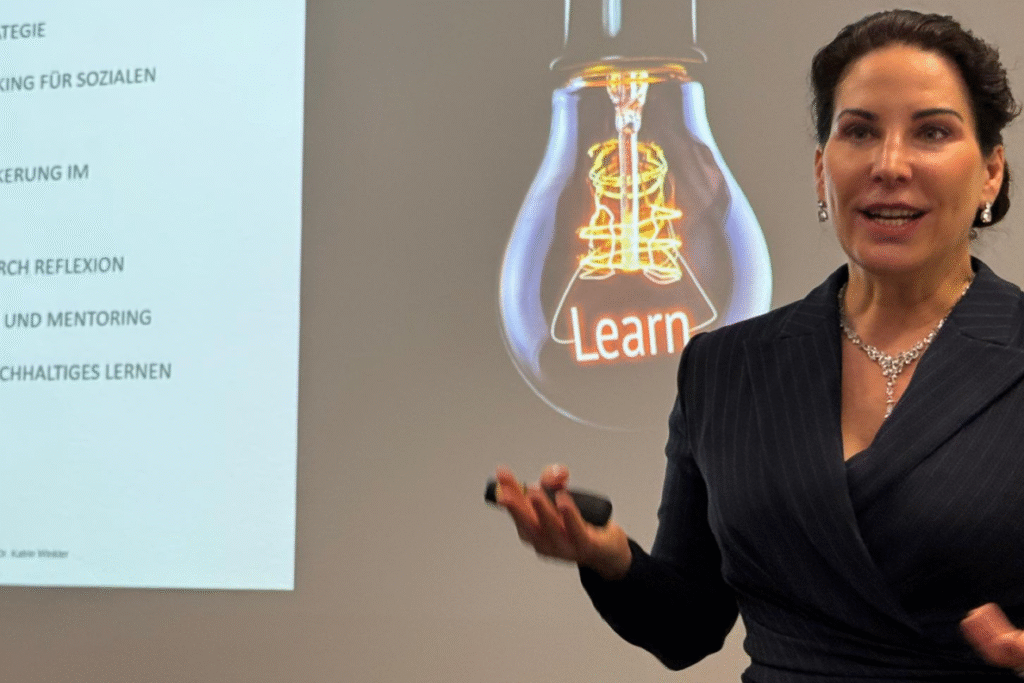 Prof. Dr. Katrin Winkler bei einem Vortrag. Sie hat einen schwarzen Nadelstreifen Blazer an. Hinter ihr ist eine Präsentationsfolie zu sehen, darauf eine Glühbirne deren Glühdraht das Wort "Learn" bildet. Sie gestikuliert und schaut geradeaus ins Publikum.