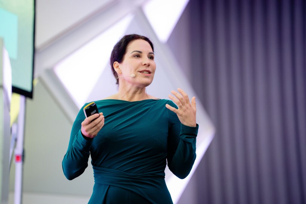 Prof. Dr. Katrin Winkler bei den Future Days von Carsten Maschmeyer. Sie hält einen Vortrag und sieht sehr energetisch und selbstbewusst aus. Sie trägt ein petrolfarbenes Kleid und hat eine Beamer-Fernbedienung in der Hand.