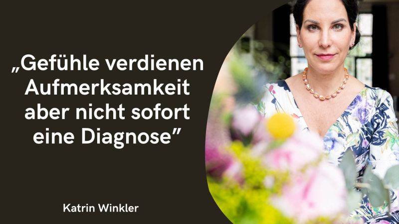 Auf der linken Seite befindet sich ein tiefdunkler, fast schwarzer Hintergrund, auf dem in markanten weißen Buchstaben das Zitat steht: „Gefühle verdienen Aufmerksamkeit aber nicht sofort eine Diagnose“. Darunter ist klein der Name Katrin Winkler platziert. Die rechte Seite zeigt die Autorin selbst in einem kreisförmigen Ausschnitt. Die Frau blickt mit einem sanften, offenen Lächeln direkt in die Kamera. Sie trägt ein helles Kleid mit blau-violettem Blumenmuster und eine Kette mit warmen, orangefarbenen Steinen. Im Vordergrund ragen unscharfe rosa und gelbe Blüten ins Bild, was dem Foto eine weiche, freundliche Tiefe verleiht. Die Gestaltung wirkt insgesamt sehr modern, ruhig und wertschätzend.