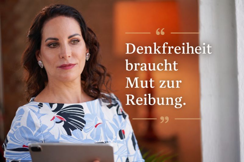 Es zeigt eine Frau (Katrin Winkler) auf der linken Seite, die nicht frontal in die Kamera blickt, sondern nachdenklich zur Seite und nach unten, während sie ein Tablet hält. Sie trägt ein Oberteil mit einem hellblau-weißen Blumen- und Grafikmuster sowie auffällige, silberne, Ohrringe. Ihr dunkles Haar ist leicht gewellt. Die rechte Bildhälfte wird von einem großen, warmen, orangefarbenen, unscharfen Hintergrund dominiert. Darüber liegt das Zitat in klaren, weißen Buchstaben mit Serifen: „Denkfreiheit braucht Mut zur Reibung.“, eingerahmt von eleganten, goldfarbenen Anführungszeichen.
